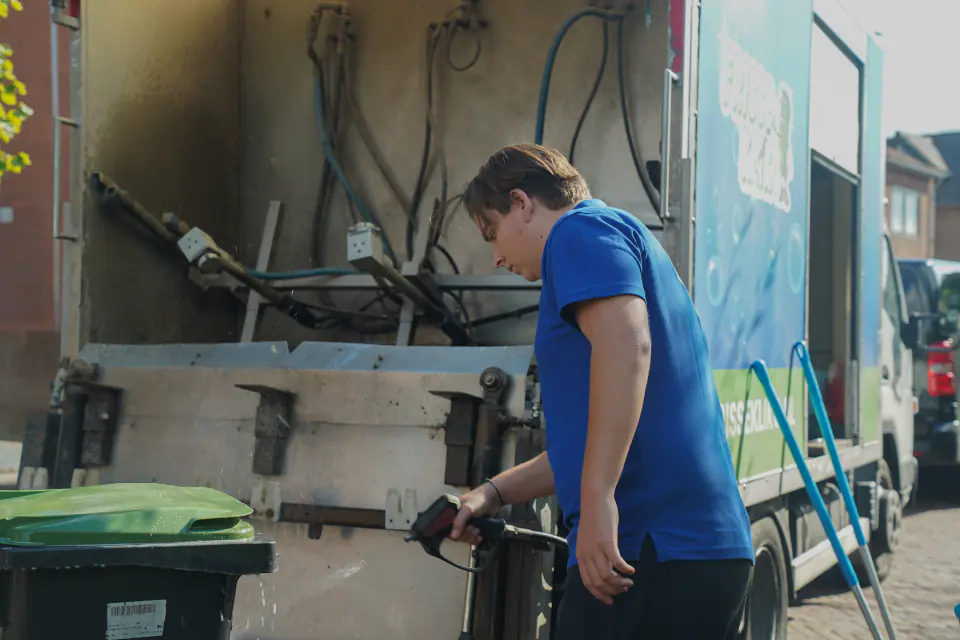 Medewerker van Frisse Kliko spuit een containerdeksel schoon bij de reinigingswagen.